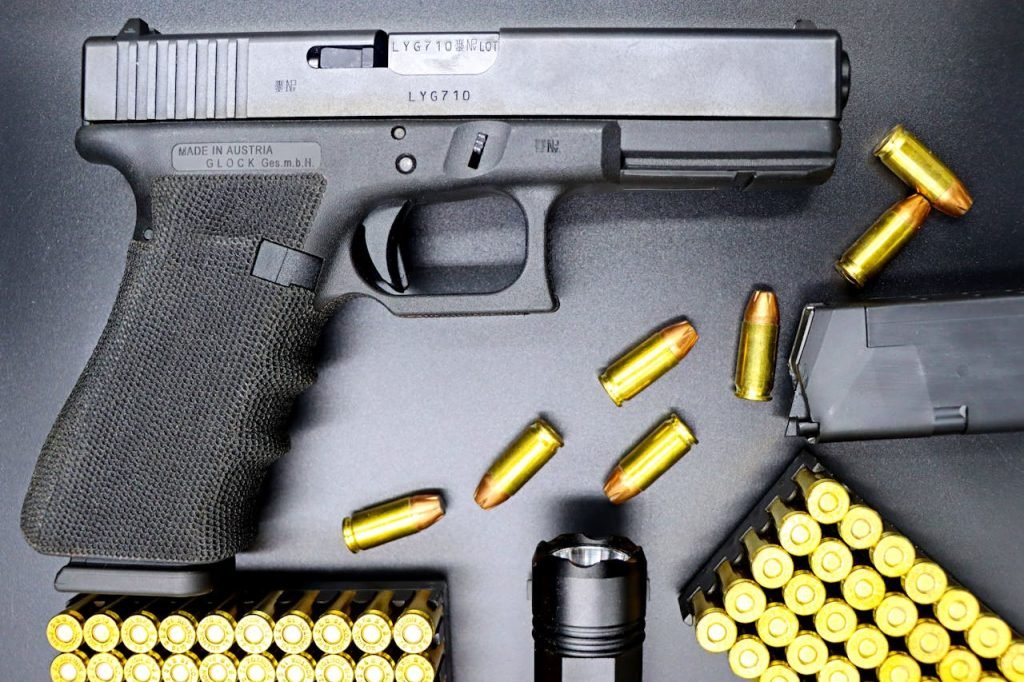 Top view of a Glock pistol, bullets, and flashlight on a black surface, showcasing firearms and ammunition.